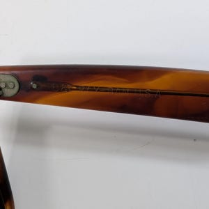 Op de afbeelding: Zonnebrilpoot met schildpadpatroon, voorzien van een metalen scharnier en de inscriptie B&L RAY-BAN U.S.A. aan de binnenzijde. Het montuur heeft een bruin en amberkleurig gemarmerd uiterlijk met een gladde, gepolijste afwerking.