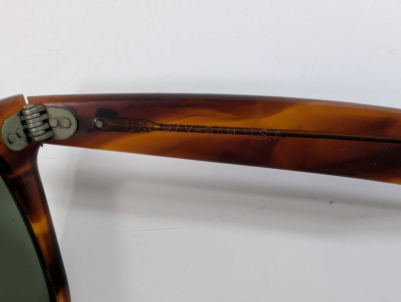 Op de afbeelding: Zonnebrilpoot met schildpadpatroon, voorzien van een metalen scharnier en de inscriptie B&L RAY-BAN U.S.A. aan de binnenzijde. Het montuur heeft een bruin en amberkleurig gemarmerd uiterlijk met een gladde, gepolijste afwerking.