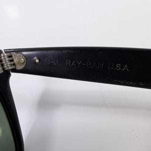 Op de afbeelding: Zwarte zonnebril met een metalen scharnier en de tekst B&L RAY-BAN U.S.A. gegraveerd aan de binnenzijde van de brilpoot. Het montuur is gemaakt van een donker materiaal met een zichtbaar scharniermechanisme dat de poot met het frame verbindt.