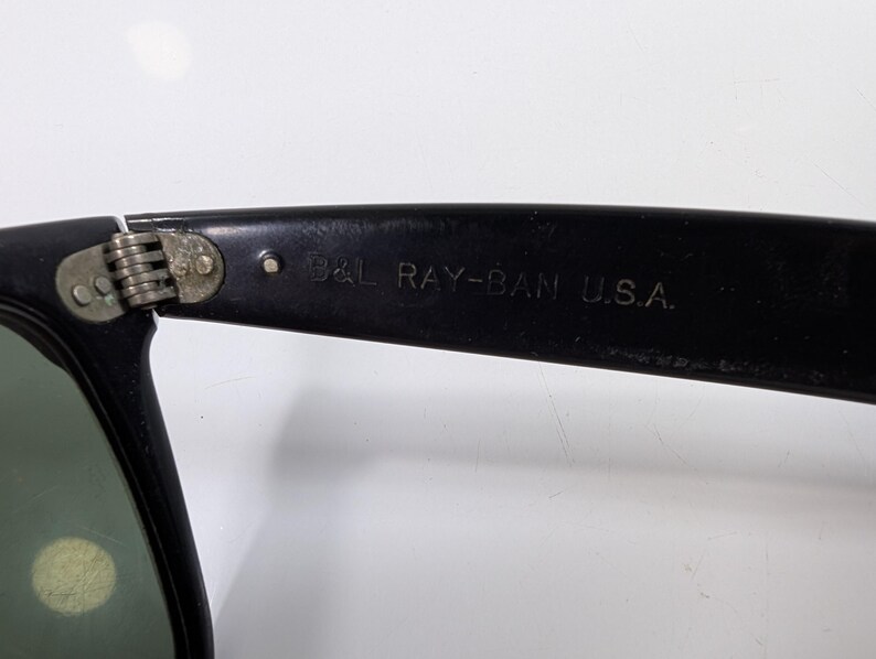 Op de afbeelding: Zwarte zonnebril met een metalen scharnier en de tekst B&L RAY-BAN U.S.A. gegraveerd aan de binnenzijde van de brilpoot. Het montuur is gemaakt van een donker materiaal met een zichtbaar scharniermechanisme dat de poot met het frame verbindt.