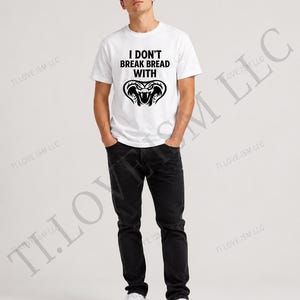 May include: A white short-sleeved t-shirt featuring the black text I DON'T BREAK BREAD WITH above a black graphic illustration of a cobra snake head. The shirt is paired with black denim jeans and white sneakers, creating a casual outfit.