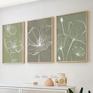May include: Three framed floral art prints. Each print features a white line drawing of a flower on a green background. The frames are made of light-colored wood. The prints are displayed on a white wall.