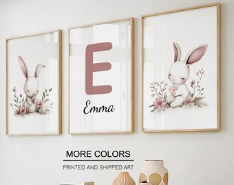Könnte beinhalten: Drei gerahmte Kunstdrucke. Einer zeigt ein Kaninchen, ein weiterer den Buchstaben "E" mit dem Namen "Emma" und der dritte ein Kaninchen mit Blumen. Die Kaninchenillustrationen sind weiß mit rosa Akzenten. Die Rahmen sind aus hellem Holz. Text: "MORE COLORS" und "PRINTED AND SHIPPED ART".