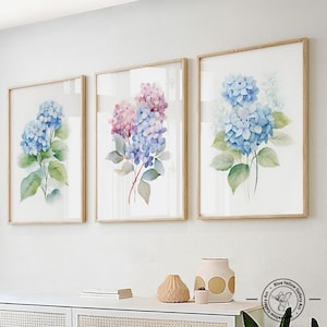 May include: Three framed watercolor paintings of hydrangeas. The paintings are in shades of blue, pink, and purple. The frames are light wood.