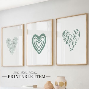 Può includere: Tre stampe d'arte da parete stampabili che presentano diversi design di cuori astratti in un colore verde salvia. Le stampe sono incorniciate in una cornice di legno chiaro e sono appese a una parete bianca.