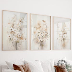 Könnte beinhalten: Drei gerahmte Kunstdrucke mit Blumenmotiven. Jeder Druck zeigt eine Aquarellmalerei von weißen und beigen Blumen mit grünen Stielen. Die Rahmen sind aus hellem Holz. Das Kunstwerk wird über einem weißen Sofa mit Kissen ausgestellt.