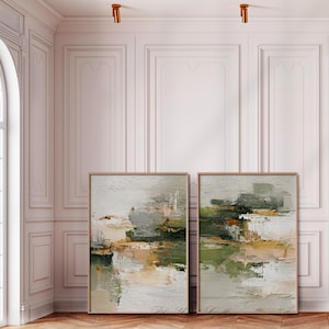 May include: Two framed abstract paintings with neutral tones. The artwork features a textured, layered effect with shades of white, gray, green, and gold. The paintings are displayed against a white wall with decorative molding, creating a gallery-like aesthetic.
