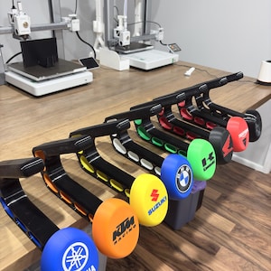 May include: A row of black motorcycle handlebar end weights featuring colourful circular caps with various brand logos including Yamaha, KTM, Suzuki, BMW, Kawasaki, Honda, and Triumph, each with matching interior colour accents on the mounting arms.