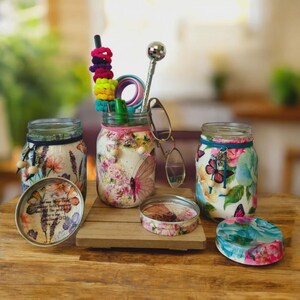 May include: Several decorated mason jars with floral and butterfly patterns. One jar contains colourful hair ties and a disco ball wand. The lids and jars feature pink, blue, and white floral designs, creating a decorative theme.