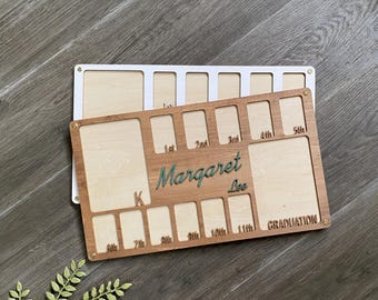 May include: Wooden photo frame with 11 slots for pictures, labeled with grades from kindergarten to 11th grade. The frame is personalized with the name "Margaret Lee" and the word "Graduation".