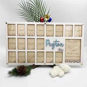 May include: White wooden photo frame with 16 slots for pictures. The frame is engraved with the words "Hello World", "1 year", "2 years", "3 years", "4 years", "Pre-K", "Kindergarten", "1st grade", "2nd grade", "3rd grade", "4th grade", "5th grade", "6th grade", "7th grade", "8th grade", "9th grade", "10th grade", "11th grade", "Class of 2039", "The days were long but the years are short", "Senior". The frame also has the name "Peyton Cha" engraved on it.