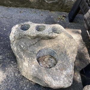 Può includere: Una grande pietra naturale grigia con tre fori scavati. La pietra ha una superficie ruvida e testurizzata e sembra essere fatta di granito.