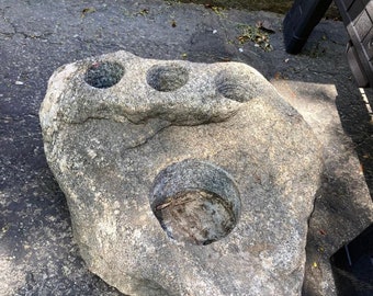 May include: A large, gray, natural stone with three carved-out holes. The stone has a rough, textured surface and appears to be made of granite.