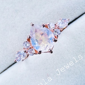 May include: Rose gold ring with a pear-cut, iridescent gemstone at its centre, encircled by smaller, clear stones. The ring is displayed on a blue velvet surface, with the text "Nebula Jewels In" visible.