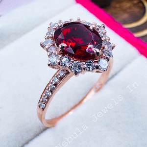 May include: Rose gold ring featuring a large, oval-cut, deep red gemstone surrounded by smaller white gemstones. The ring's band is also adorned with small, clear stones. The ring is displayed in a white jewelry box with a pink ribbon.