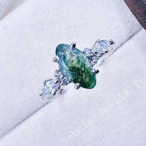 May include: A silver ring featuring a large, oval-shaped green moss agate gemstone, accented with smaller clear gemstones. The ring is set on a white surface with the text "Nebula Jewels In".