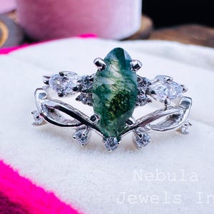 Könnte beinhalten: Ein silberner Ring mit einem grün-moos-achat-Edelstein im Marquise-Schliff und kleinen, klaren Akzentsteinen. Der Ring liegt auf einer weißen Oberfläche mit dem Text "Nebula Jewels In".