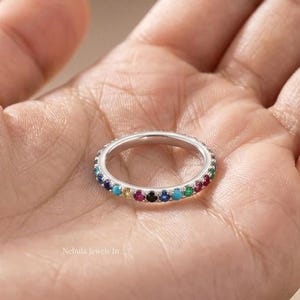 May include: A silver-toned band ring featuring a continuous row of small, round, multi-coloured gemstones in shades of red, orange, yellow, green, blue, and purple, set evenly around the entire circumference of the metal band.