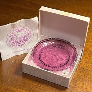 Puede incluir: Un plato de cristal rosa con borde festoneado está en una caja blanca. El plato tiene un diseño grabado y es una edición limitada de 1987. Se ve un papel con texto.