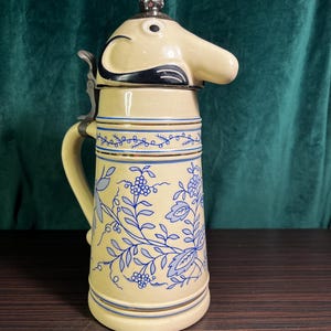 以下が含まれることがあります： クリーム色の陶器製ビアジョッキで、青い花柄が施されています。蓋は長い鼻を持つ象の頭を模した金属製で、金属製のハンドルとヒンジ付きの蓋、装飾的な金属製のつまみが付いています。