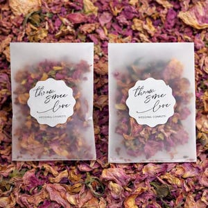 Puede incluir: Dos pequeños paquetes translúcidos llenos de pétalos de flores secas, con la etiqueta "throw some love WEDDING CONFETTI". Los paquetes están rodeados de un lecho de pétalos secos en tonos rosas, morados y marrones, perfectos para una boda.