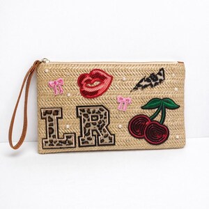 Puede incluir: Un clutch tejido beige con una correa de muñeca marrón. El clutch está decorado con labios rojos con lentejuelas, letras "LR" con estampado de leopardo, lazos rosas, un rayo con estampado de leopardo y cerezas. Pequeñas perlas están dispersas.