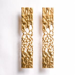 May include: A pair of gold-toned earrings with a textured, openwork design. The earrings are rectangular in shape and have a slightly irregular, organic look.