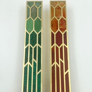May include: Two rectangular cabinet pulls with brass frames. One features green malachite and the other red agate, both with geometric designs. These decorative pulls are ideal for adding a touch of elegance to furniture.