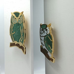 Może przedstawiać: Dwa dekoracyjne uchwyty do szafek w kształcie sowy. Sowy wykonane są z zielonego i białego kamienia, ze złotymi metalowymi akcentami. Siedzą na brązowej gałęzi. Uchwyty są zamontowane na białej powierzchni.