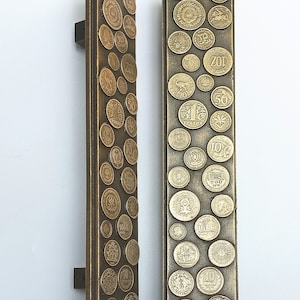 May include: Two bronze door pulls, each featuring a rectangular design adorned with various vintage coins. The coins are of different sizes and denominations, adding a unique, antique aesthetic to the hardware.