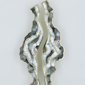 May include: Two abstract wall art pieces with a wavy design. The pieces feature a mosaic of black, grey, and white iridescent shell fragments, outlined with a gold-coloured metal. The central section is a smooth, gold-coloured metal.