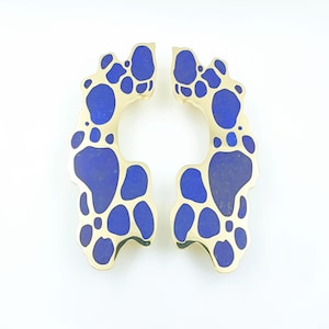 May include: A pair of gold-toned earrings with a unique, organic design. Each earring features irregular shapes filled with deep blue material, creating a striking contrast. The earrings have a curved, open shape.