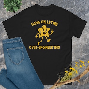 Könnte beinhalten: Ein schwarzes Kurzarm-T-Shirt mit einer gelben Grafik eines lächelnden Sterns mit Armen und Beinen. Der Text über dem Stern lautet HANG ON, LET ME und der Text unter dem Stern lautet OVER-ENGINEER THIS in einer fetten gelben Schrift.