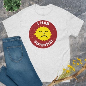 May include: A light gray t-shirt with the text "I HAD POTENTIAL" in a distressed red circle. The circle contains a yellow sun with a sad face. Blue jeans are folded below the shirt.
