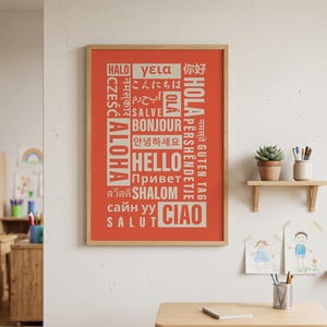 Op de afbeelding: Een rechthoekige wandkunstprint in een lichthouten lijst met een levendige oranje achtergrond waarop het woord Hallo in verschillende talen en schriften staat, waaronder Engels, Frans, Spaans, Italiaans, Duits en Japans in het wit.
