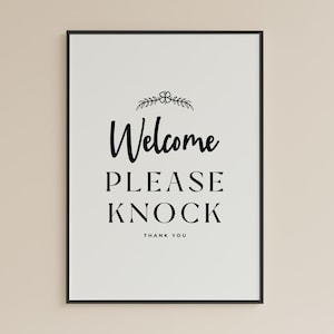 Puede incluir: Una impresión enmarcada con las palabras "Welcome PLEASE KNOCK THANK YOU" en escritura negra y letras de bloque. La impresión tiene un fondo blanco y un marco negro, con un pequeño diseño floral encima de la palabra "Welcome".