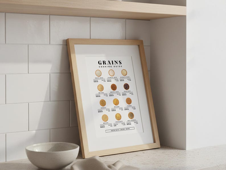 Może przedstawiać: Oprawiony plakat "GRAINS COOKING GUIDE" z ilustracjami r&oacute;żnych zb&oacute;ż. Plakat jest w jasnej drewnianej ramie, oparty o białą kafelkową ścianę. Biała ceramiczna miska i beżowa szmatka na pierwszym planie.