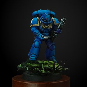 May include: A blue miniature figurine of a space marine, holding a black rifle. The figurine has gold accents and a skull emblem. It stands on a base with green foliage, set against a dark background.