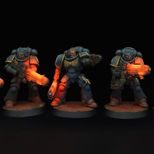 May include: Five miniature Space Marine figures in dark blue and gold armour, each holding a glowing orange weapon. The figures stand on brown bases against a dark background. The figures are posed as if in action.