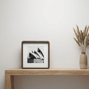 May include: A framed black and white photograph of the Sydney Opera House. The square picture is in a dark wood frame, resting on a light wood shelf. A vase with dried plants is to the right of the frame.