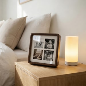 May include: A dark wood picture frame displaying four monochrome photographs. The frame is square with curved edges. Beside it, a small cylindrical lamp with a white base emits a warm, soft light. The items are on a wooden bedside table.