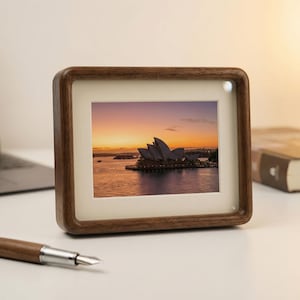 May include: A square wooden photo frame displaying a sunset view of the Sydney Opera House. The frame is dark brown with a cream mount. A pen lies on the white surface in front of the frame, with a book in the background.