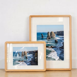 May include: Two framed landscape prints depicting a coastal scene with rock formations in the sea. The frames are made of light-coloured wood. The prints show a blue ocean and sky, with a simple, modern design.