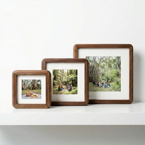May include: Three wooden picture frames of different sizes with rounded corners. Each frame has a photograph with a white mount. The frames are displayed on a white shelf. The photos show people in a forest setting.