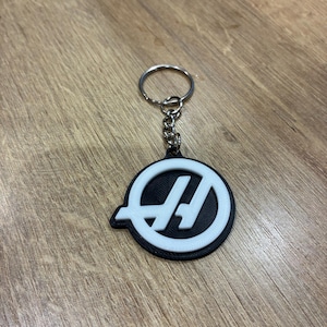 Puede incluir: Un llavero en blanco y negro con un emblema circular con un diseño estilizado de la letra "H". El llavero tiene un anillo y una cadena plateados, y el emblema está sobre un fondo negro.