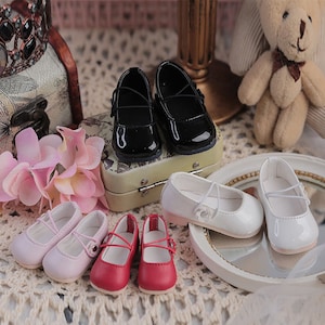 May include: Six pairs of doll shoes in various colours, including black, white, pink, and red. The shoes are all closed-toe and have a strap across the instep.