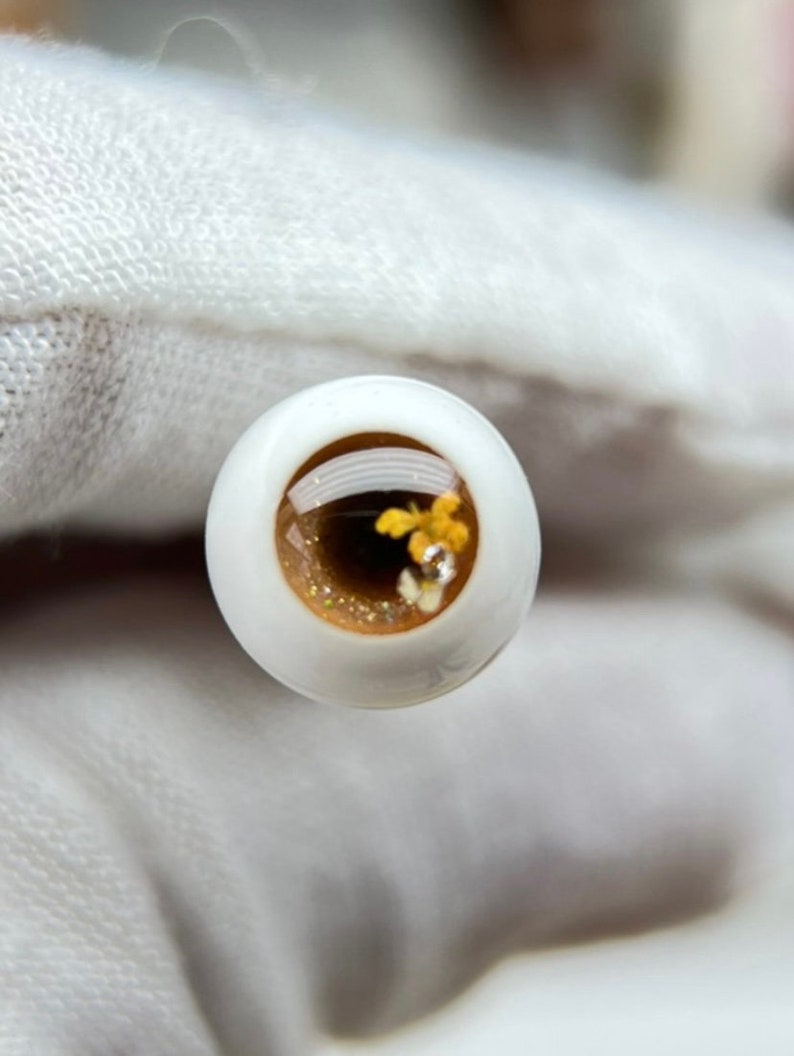 Puede incluir: Primer plano de un ojo de vidrio marr&oacute;n con purpurina dorada y peque&ntilde;as flores amarillas en su interior. El ojo est&aacute; rodeado de un fondo blanco.