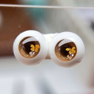 Puede incluir: Un par de ojos de mu&ntilde;eca marrones y dorados con un acabado brillante y detalles de flores amarillas.