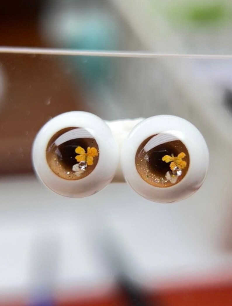 Puede incluir: Un par de ojos de mu&ntilde;eca marrones y dorados con un acabado brillante y detalles de flores amarillas.