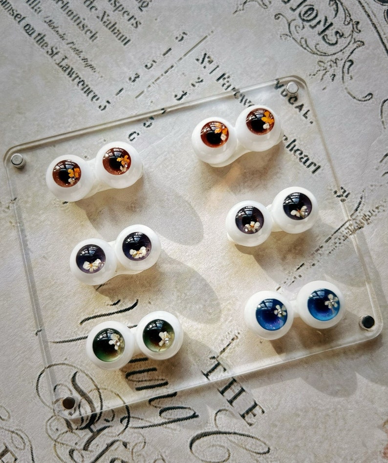 Puede incluir: Un conjunto de seis pares de ojos de mu&ntilde;eca con iris de diferentes colores y dise&ntilde;os de flores. Los ojos se muestran en una l&aacute;mina de acr&iacute;lico transparente con fondo blanco.
